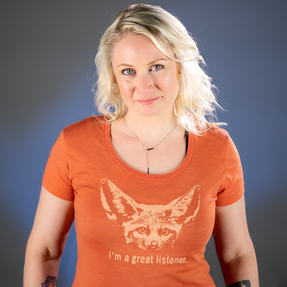 Justine Mastin color portrait. Justine is a white woman with blonde hair and is wearing an orange t-shirt with a fox on it that says I am a great listener. She is smiling and has her arms by her sides.