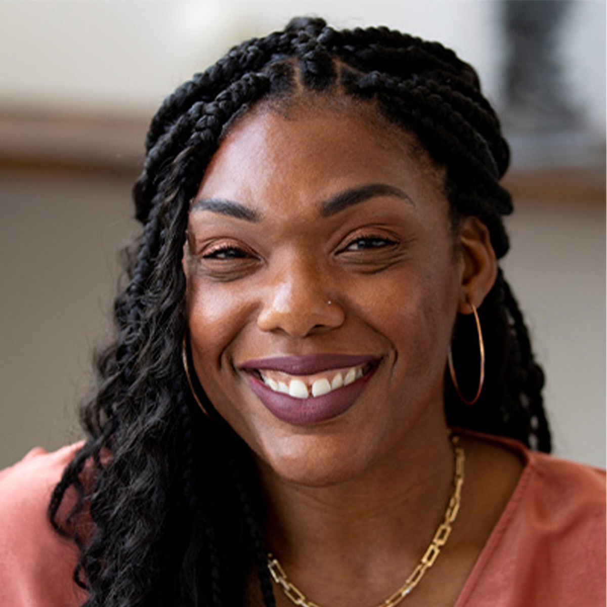 Minaa B. color portrait. Minaa B. is a Black woman who is smiling, has long braided hair, and is wearing a rose-colored wrap top, hoop earrings, and a gold necklace.