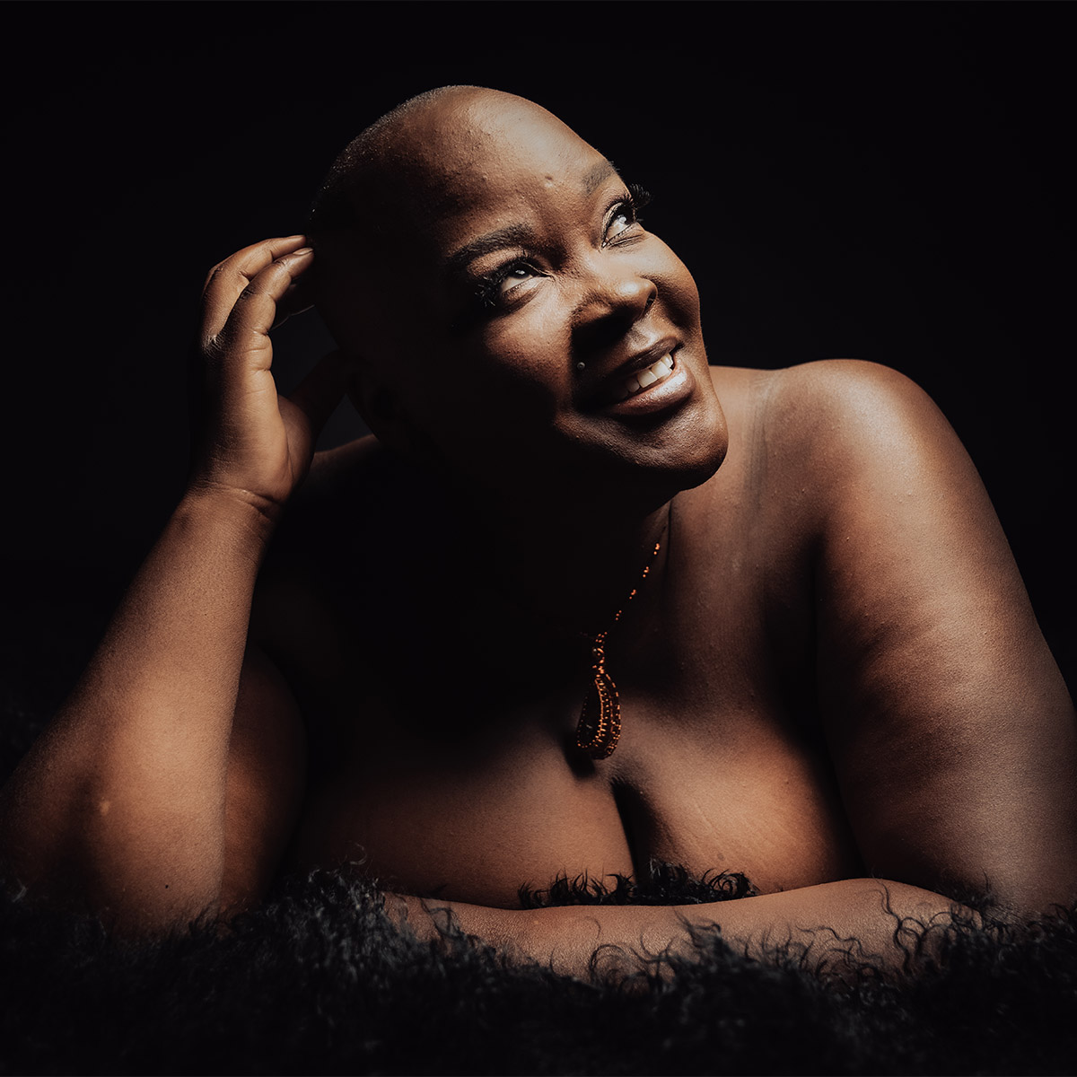 Sonya Renee Taylor in a color portrait. Sonya is a Black woman and is looking to the right and smiling. Sonya is resting on a dark, fuzzy carpet and the background is black behind her.