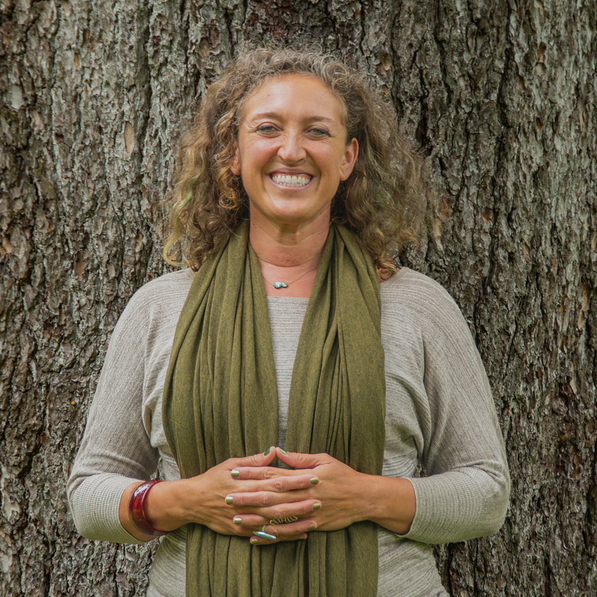 Zara Zimbardo color portrait. Zara is a woman with light colored curly hair. She is smiling, standing in front of a wide-trunk of a tree. She has her hands laced and rested on her stomach.