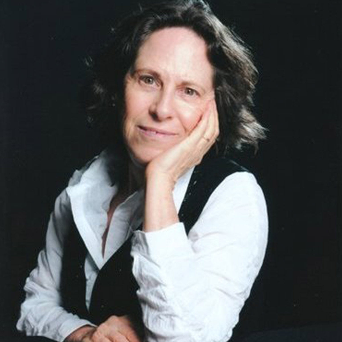 Connie Zweig color portrait. Connie is an older, white woman with dark hair that ends before her shoulders. She is wearing a white top and a black vest over it. Connie smiles and has her elbow resting on the table and her hand resting on her cheek