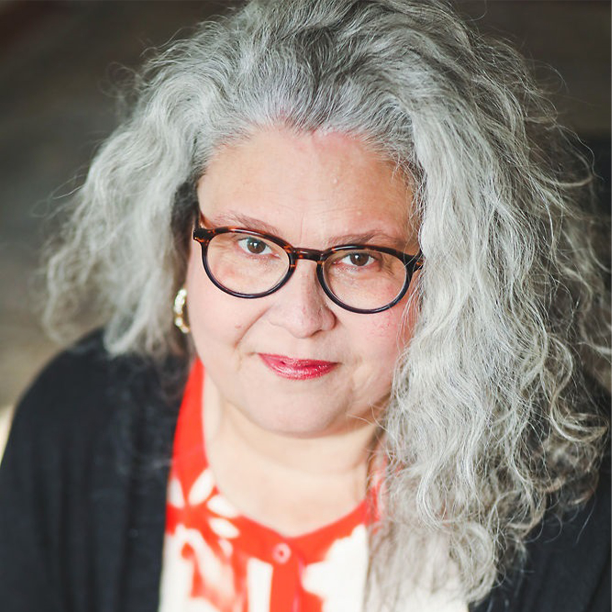 Laura Rowe color portrait. Laura has silver, gray curly hair that ends past her shoulders. She is smiling and wearing a red and white patterned top, a black cardigan and plastic, circular eyeglasses on.