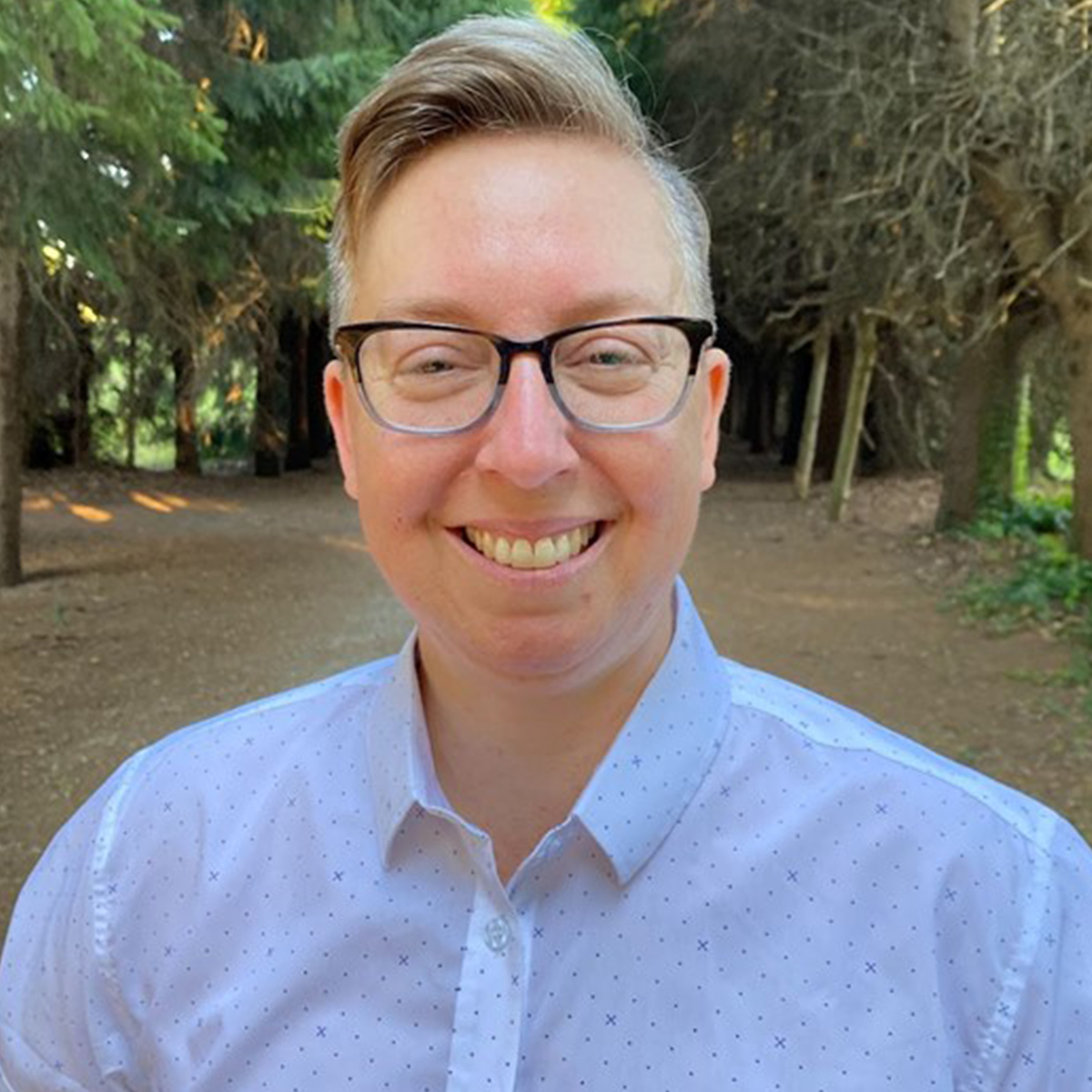 Nyck Walsh color portrait. Nyck Nyck’s identities include white, Autistic, queer, trans, and anti-Zionist Jewish. He is posed outside and is wearing two-toned glasses, a white button-down, and is smiling amongst the trees.