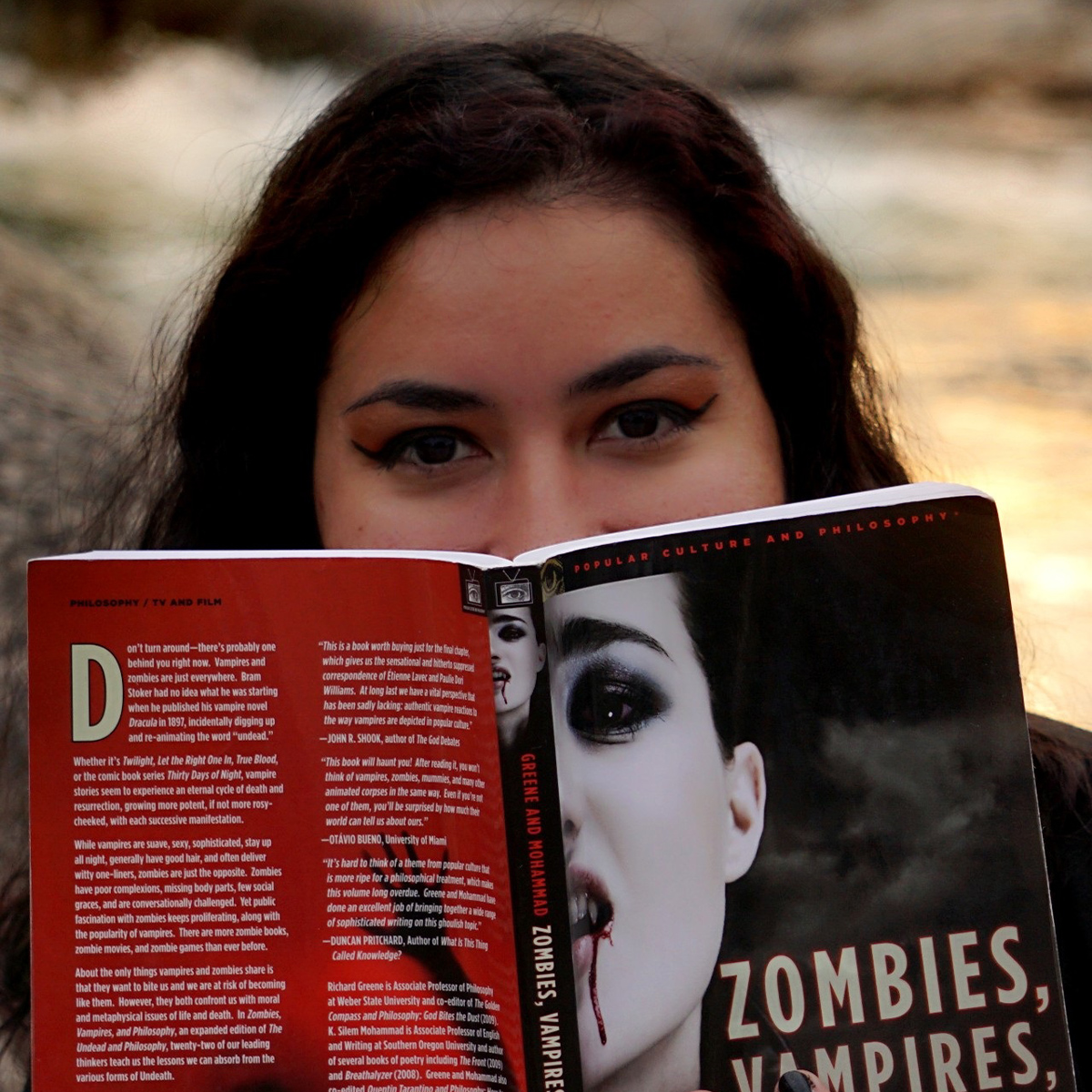 Chicana holding a book in front of the lower half of their face. They are wearing a black t-shirt with a Halloween jack o'lantern design, and have on orange eye shadow with black eyeliner make-up. The book is titled "Zombies, Vampires, and Philosophy" and there is a blurred river in the background.