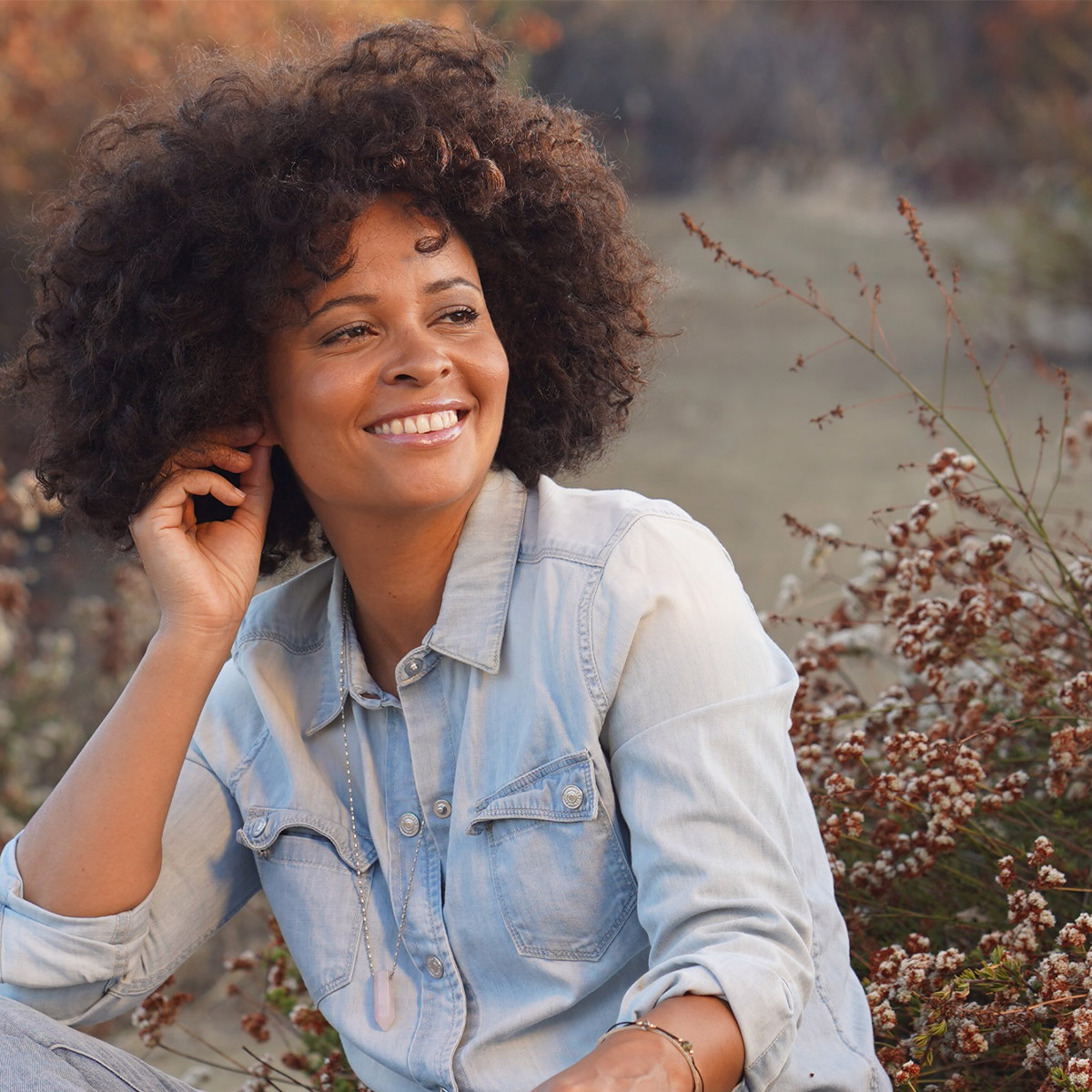 Susan Ateh color portrait. Susan is a mixed race woman who is seated cross-legged on the ground surrounded by grass and dried wildflowers. Susan has dark brown curly hair. She is smiling and has one hand resting on her ear and is wearing blue jean button-down.
