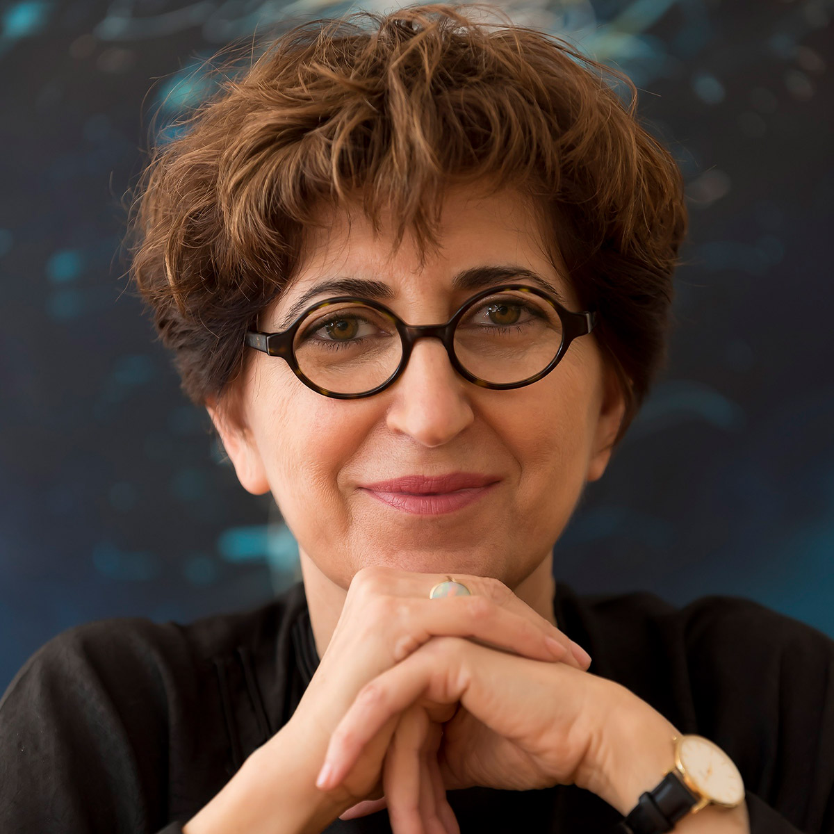 Fariba Bogzaran color portrait. Fariba is posed with both hands resting on each other and on her chin. She has short brown hair, circular eye-glasses, and a dark-colored top.