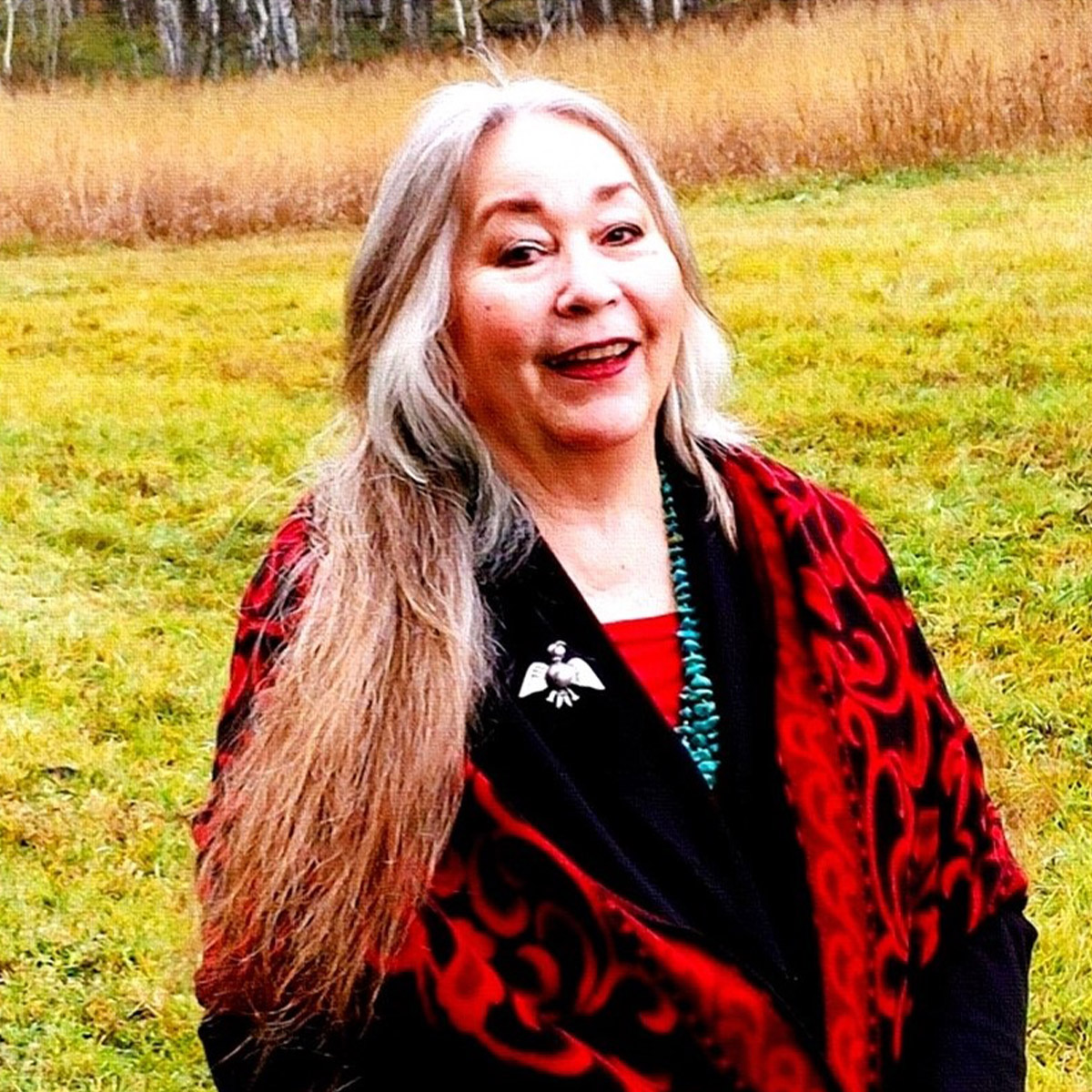 Francesca Mason Boring color portrait. Francesca is a bicultural Western Shoshone woman enrolled with the Shoshone Paiute Tribes. She is smiling, posed outside with grass and dried plants behind her. She is wearing a bright-red printed knit coat, a silver pin shaped liked a bird, and has long silver hair with brown highlights.