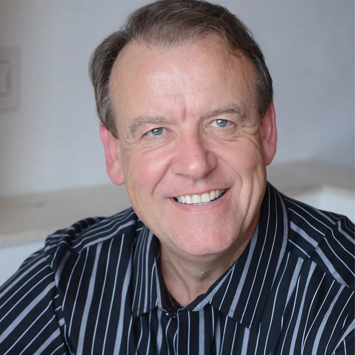 Dawson Church color portrait. Dawson is an older, white man who is smiling, posed in front of a neutral background and is wearing a vertical striped button-down shirt.