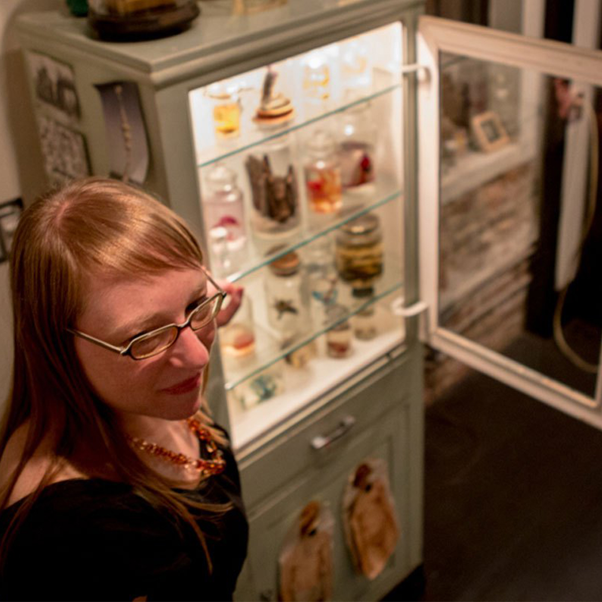 Joanna Ebenstein color portrait taken from above. Joanna is a white woman with strawberry blonde hair and is wearing glasses. A white cabinet is open and you can see glass jars inside.