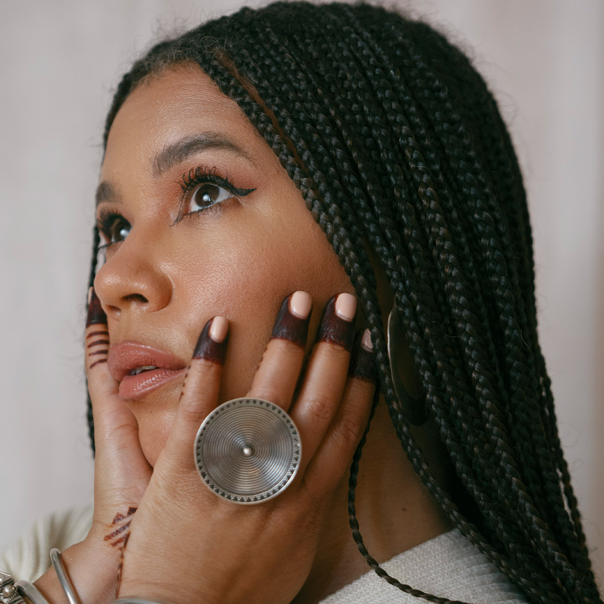 Maryam Hasnaa color portrait. Maryam is a Black woman with long, braided hair to her lower back. Her eyes are closed and her arms are raised and hands open across her chin and head.