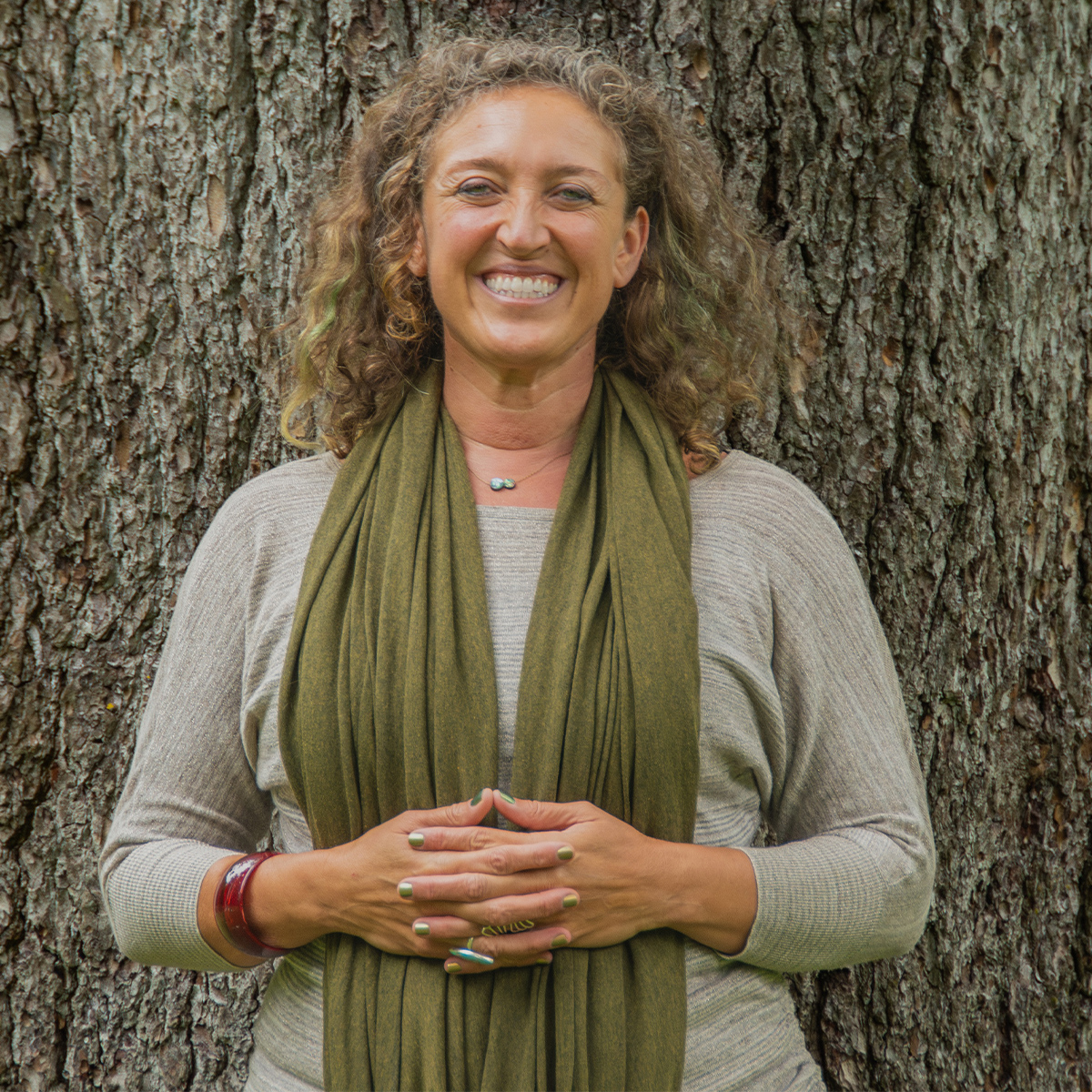 Zara Zimbardo color portrait. Zara is a woman with light colored curly hair. She is smiling, standing in front of a wide-trunk of a tree. She has her hands laced and rested on her stomach.