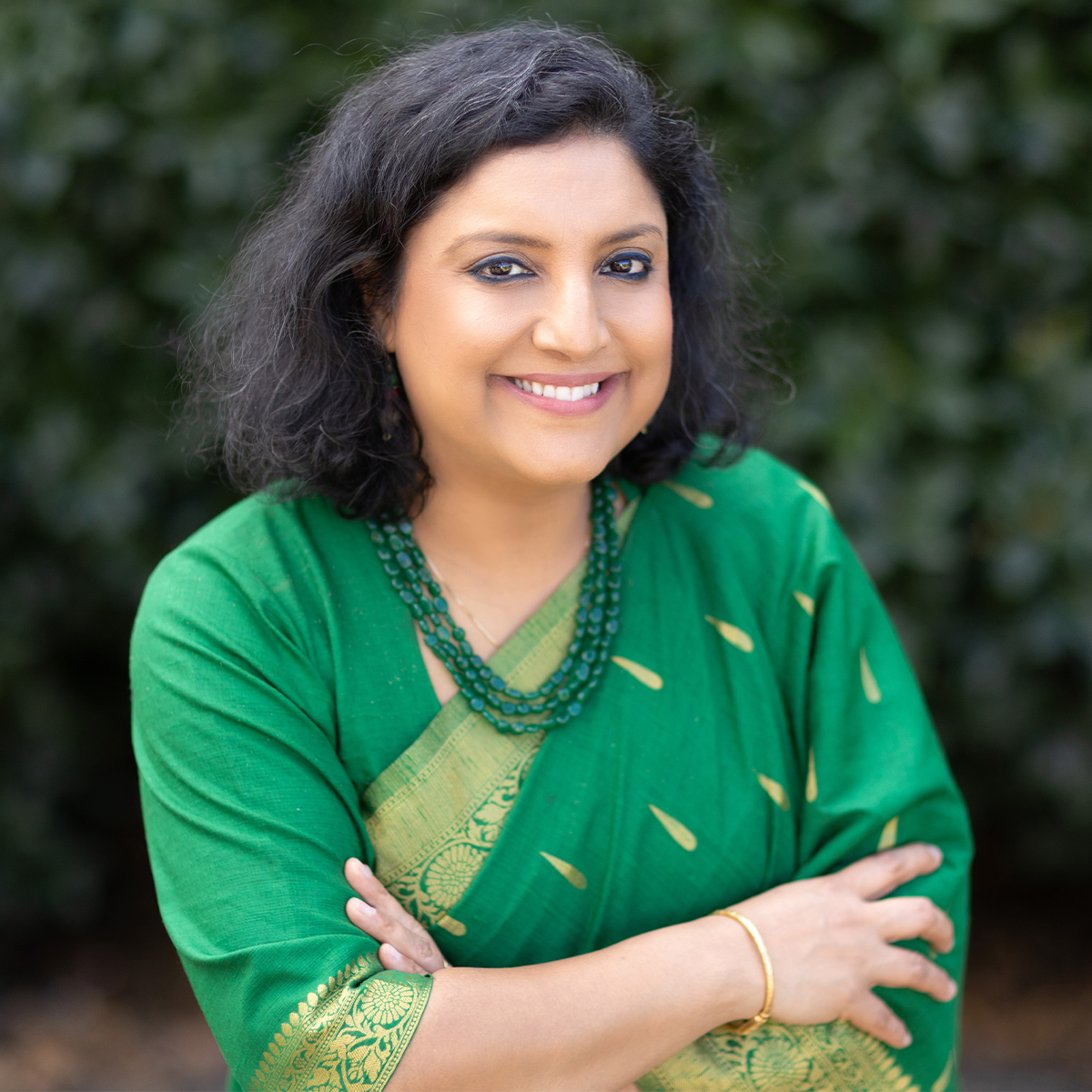 Geetanjali Chakraborty color portrait. Geetanjali has light brown skin, and shoulder length dark brown hair. She is smiling and is wearing a three strand beaded necklace and is wearing a green and gold sari. She is smiling while posing with her arms crossed.