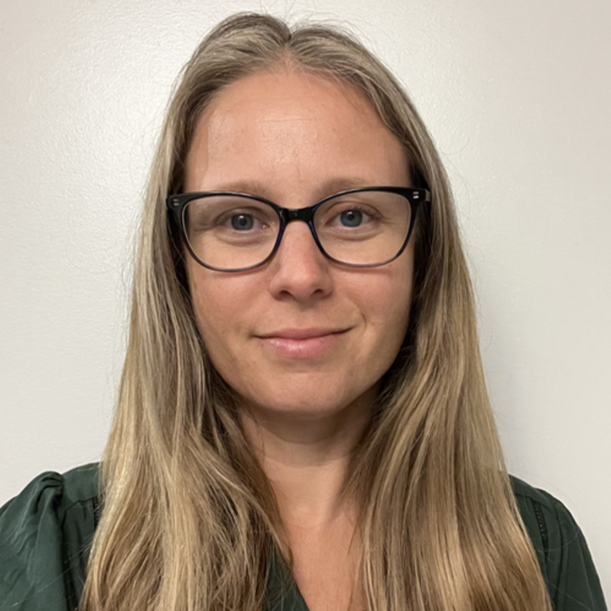 Michelle Glowa color portrait. Michelle is a white woman with long blonde hair and is wearing wide-rimmed dark-colored glasses. She is smiling in the portrait, is posed in front of a neutral wall, and is wearing a dark green colored top.