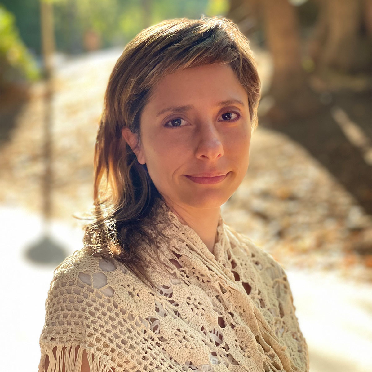 Sara Granovetter color portrait. Sara is a white woman with her hair swept to one side. She is smiling, posed outside, and is wearing a cream knit, lace-line shawl on her shoulders.