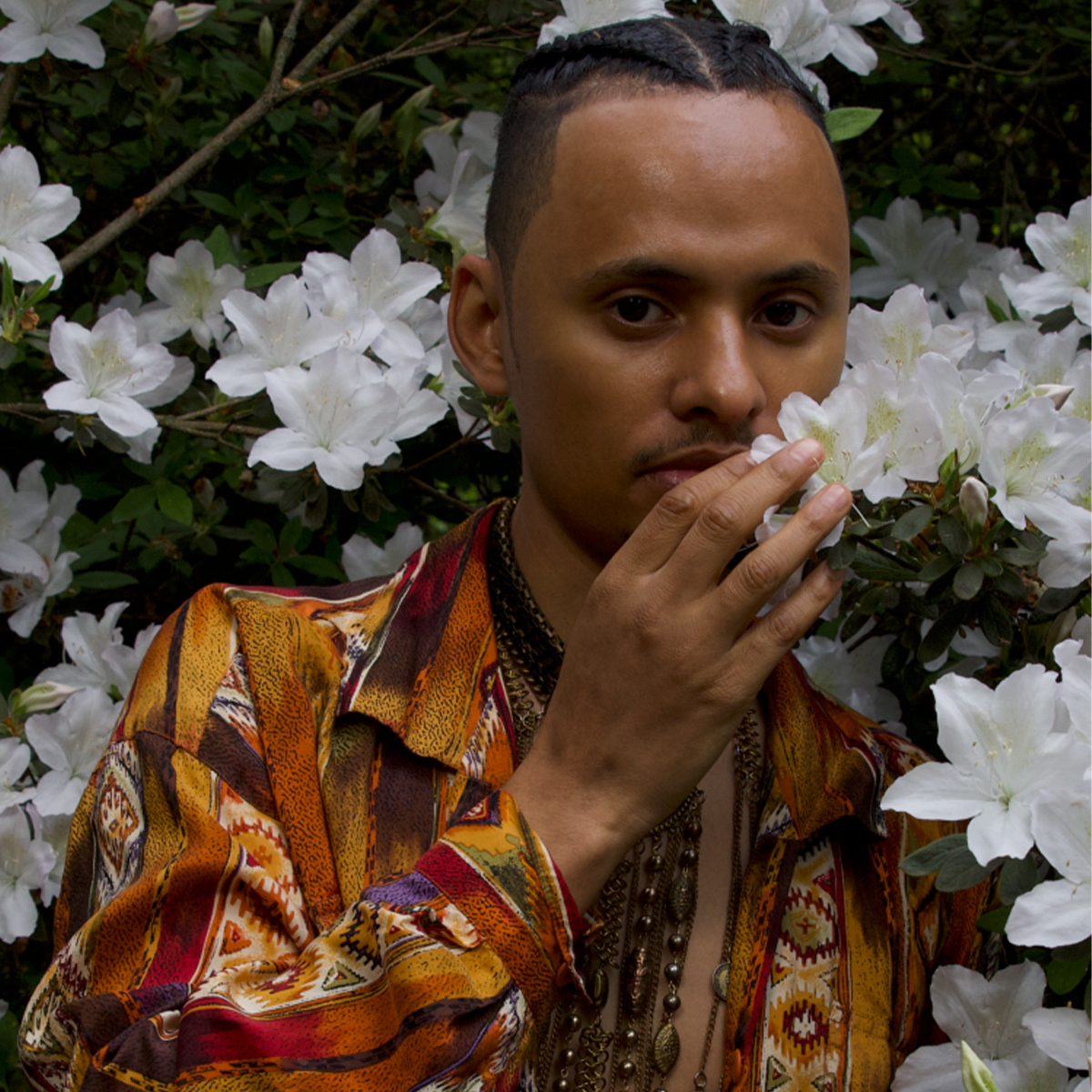 Langston Kahn color portrait. Langston is posed surrounded by white flowers from a tree or bus and is holding one close to his face. Langston is a Black, Queer, New York City-based person with his hair braded back. He is wearing a printed top with shades of brown, orange, and white.