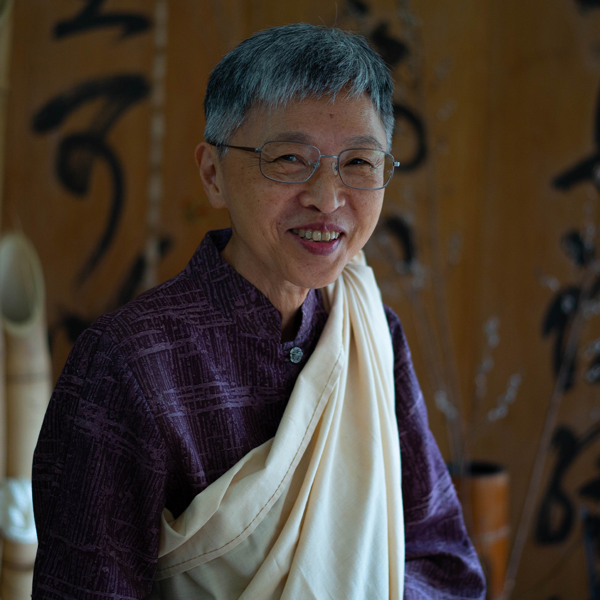 Norma Wong color portrait. Norma is Native Hawaiian and Hakka life-long resident of Hawai’i. Norma is smiling in this portrait and is posed in front of panels that have Japanese writing on them. Norma is wearing a light-colored fabric draped from one side to one shoulder and a dark colored plaid-esque printed top. 