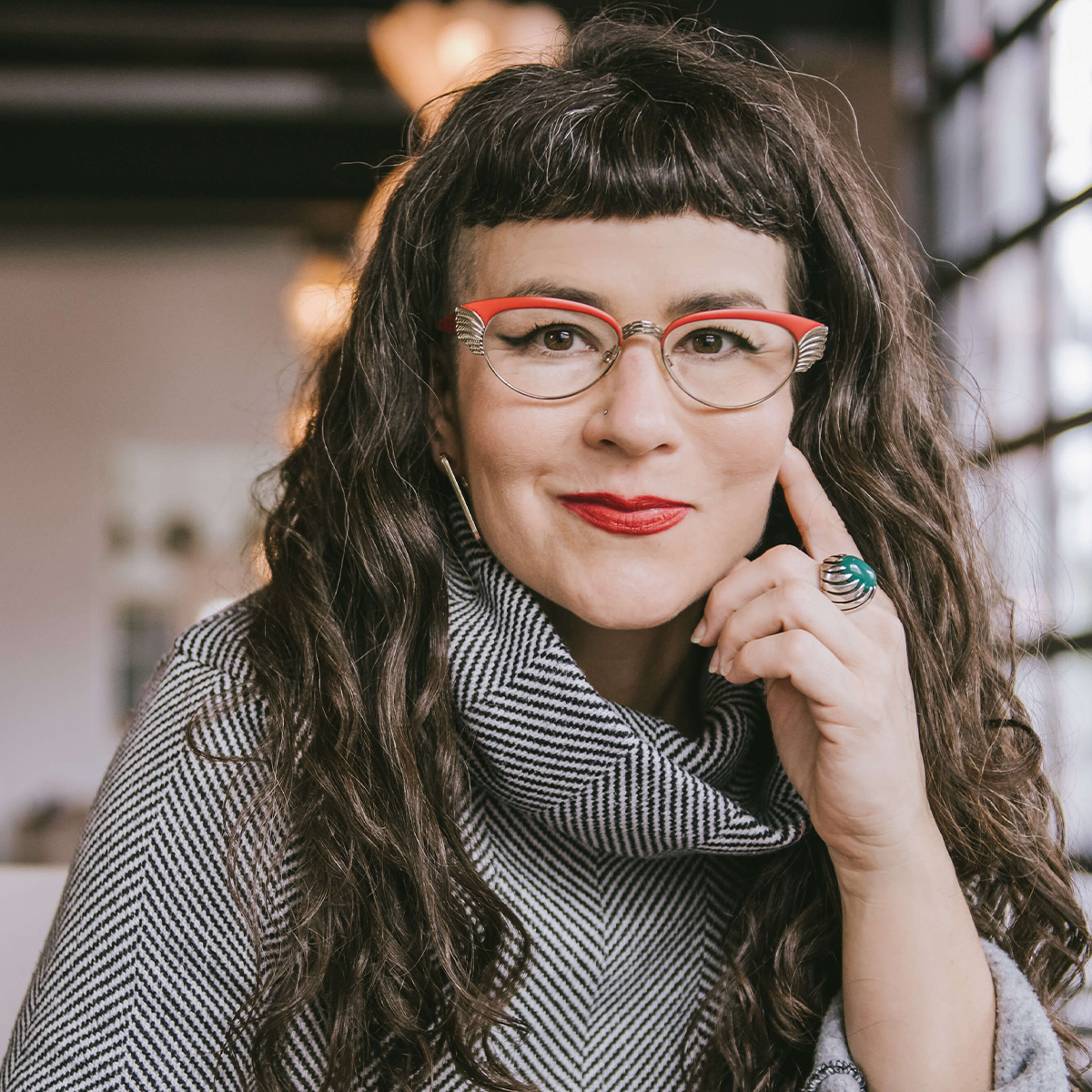 Beatriz Albina color portrait. Beatriz is a white Latina woman with long curly, brown hair that ends well past her shoulders. She is wearing red-rimmed glasses and has one hand resting on her cheek as she smiles. She is posed inside and is wearing a tweed-like top and a ring on the pointer finger that rests on her cheek.