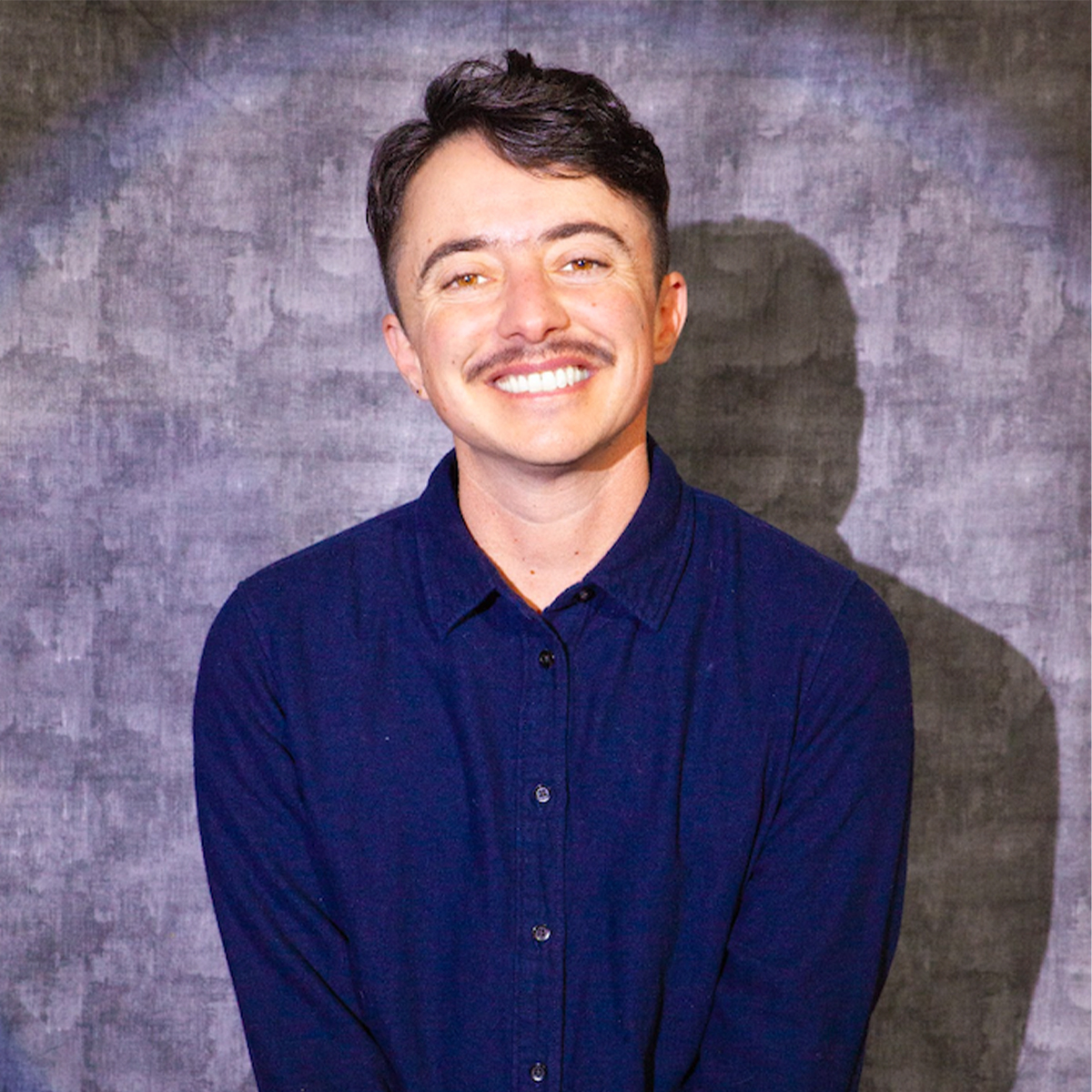 Dean Spade color portrait. Dean Spade is a white American, specifically of Jewish ethnicity; he is openly trans. He is smiling and posed in front of a light blue cloth and is seated on a stool wearing a dark-blue button-down.