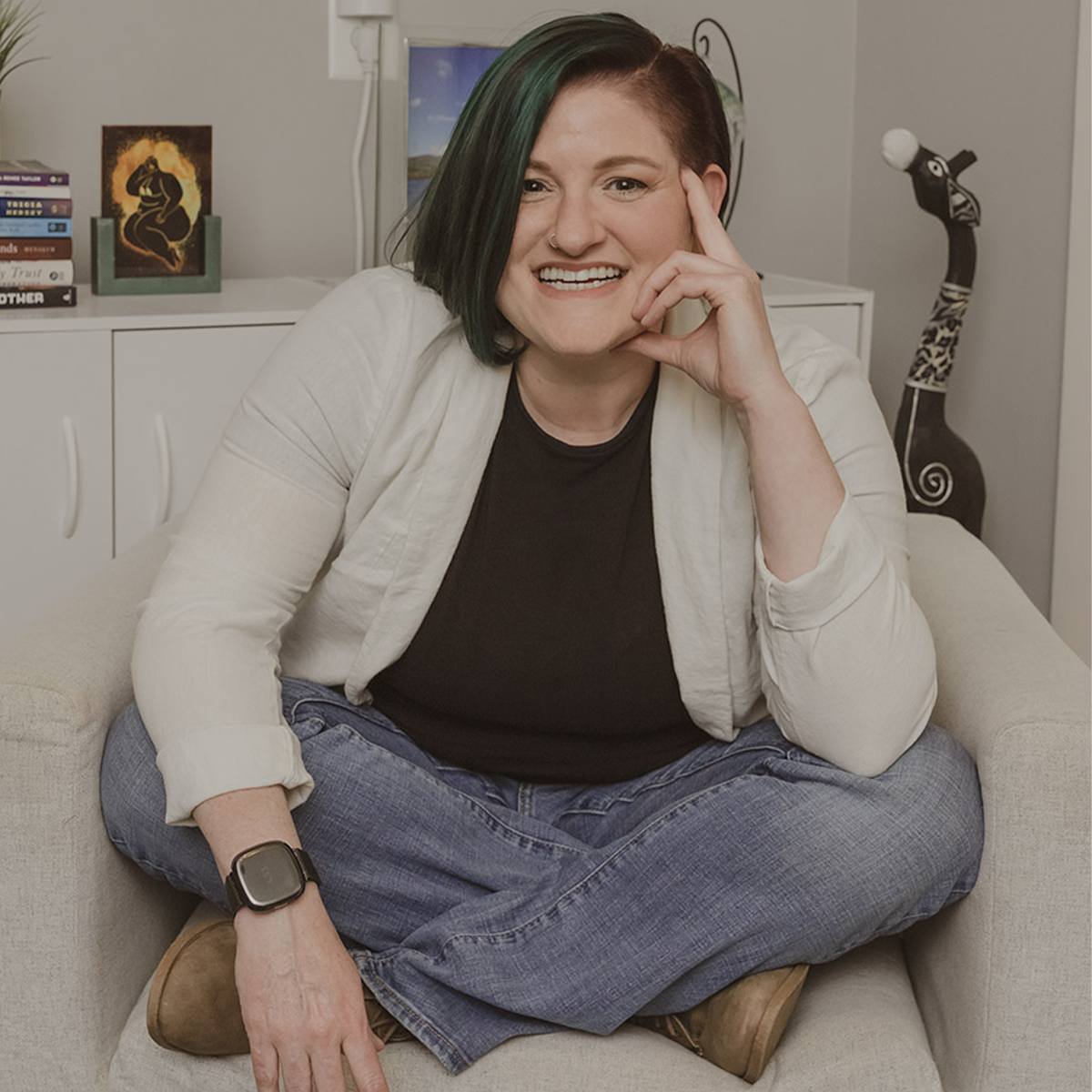 addyson tucker color portrait. addyson is genderqueer, white, small fat, and neurodivergent person who is seated cross-legged on a cream colored arm chair inside a room and has one hand resting on their cheek as they smile and the one rests on their knee.