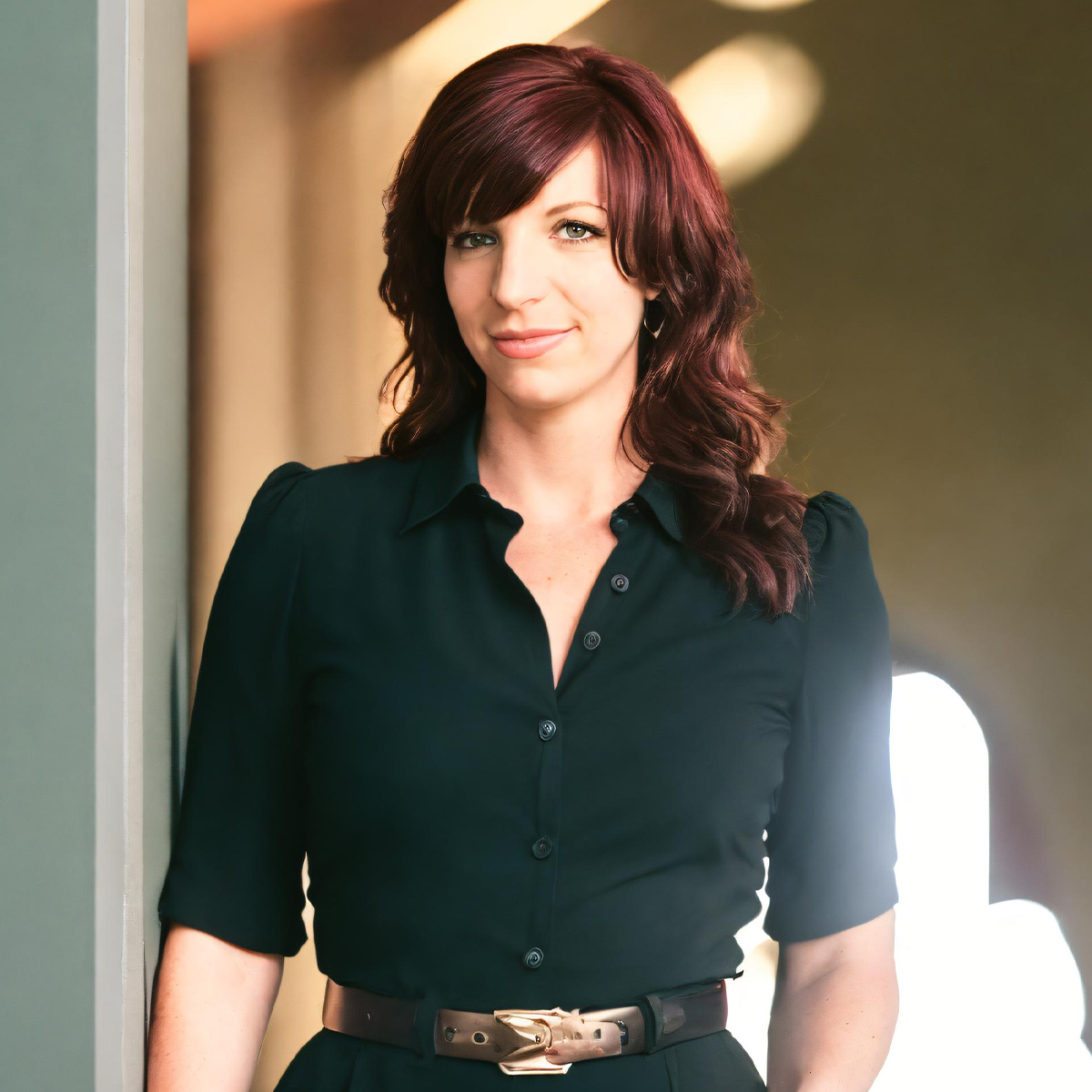 Dr. Alison Ash in a black dress with a belt standing in a doorway. Dr. Ash is a white woman with long wavy red hair that ends past her shoulders.