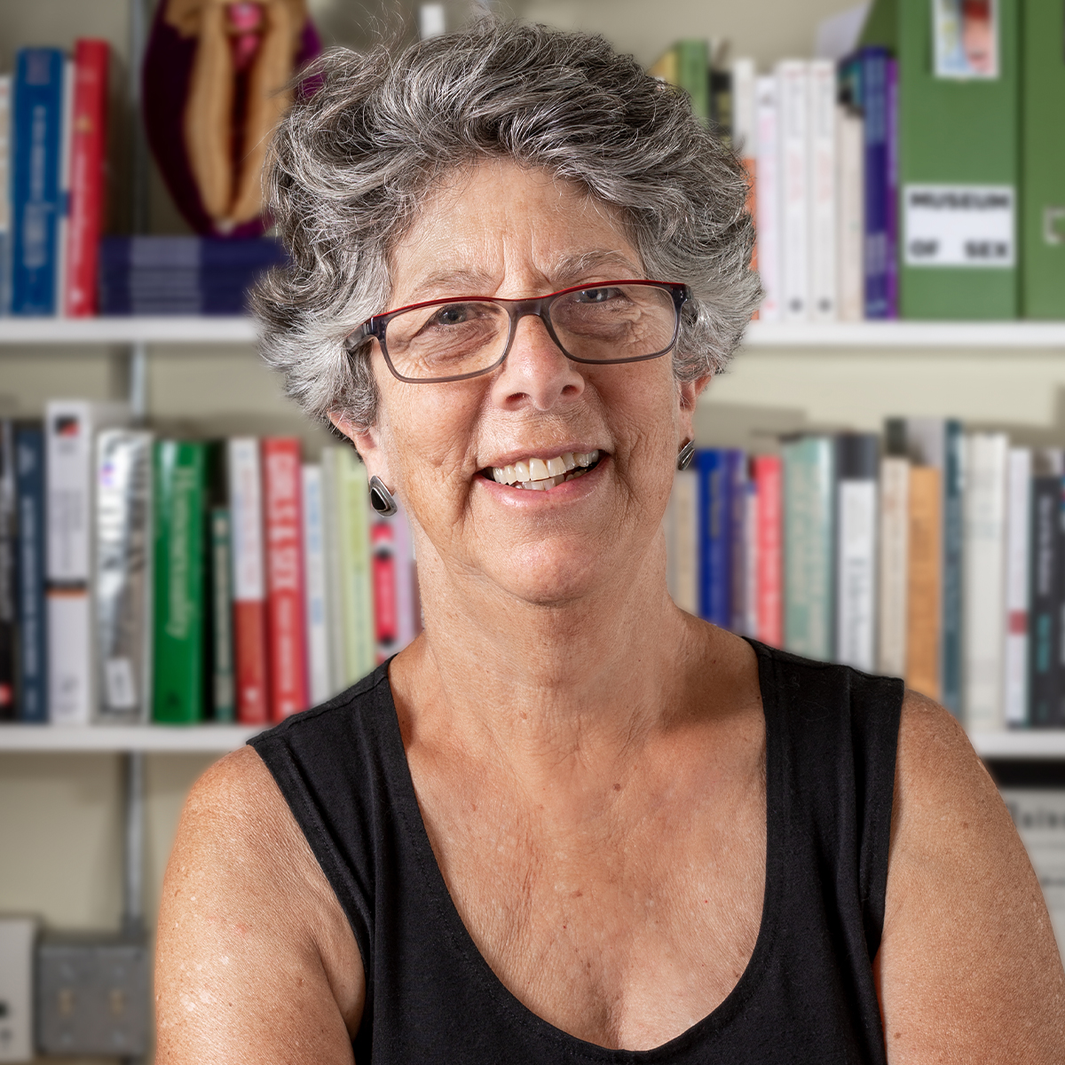 Jane Fleishman color portrait. Jane is an older, white woman with short silver/gray hair and is wearing glasses, a black sleeveless top, and is posed in front of a bookshelf.