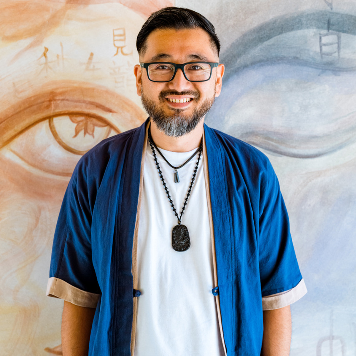 Kazu Haga color portrait. Kazu is a Asian American person who is smiling, while posed standing in front of a mural that is shades of light brown with an open eye painted on the left and shades of blue with a eye closed on the right side.