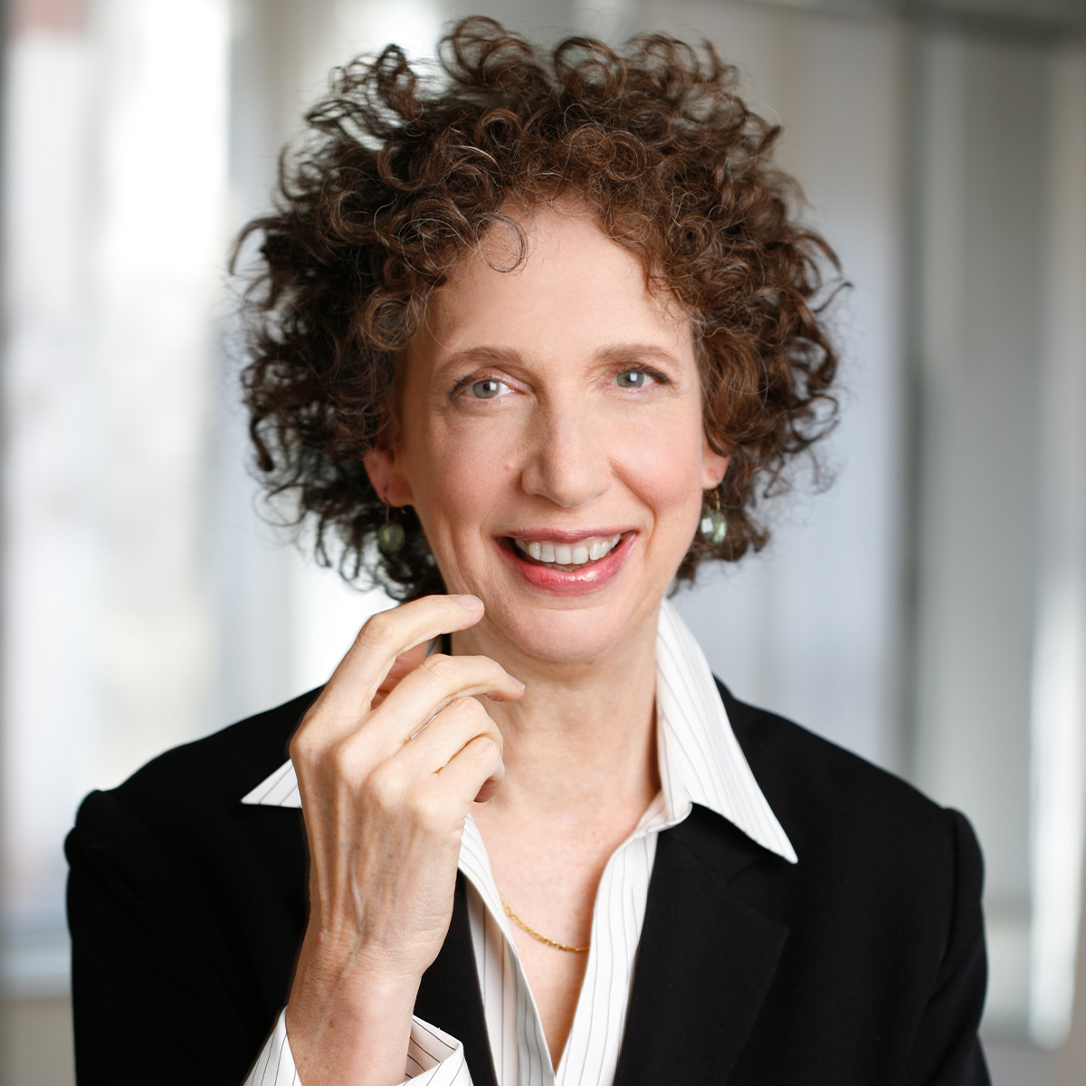 Betsy Polatin color portrait. Betsy has chin length curly reddish brown hair and is a white woman with one hand gesturing towards her face as she smiles. She is wearing a black blazer and white collared shirt.