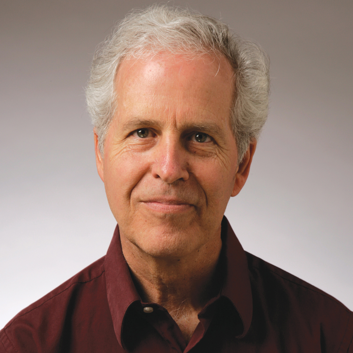 John Prendergast color portrait. John is posed in front of a neutral gradient gray background and is a white, older man with short, silver hair. He is smiling and wears a dark maroon button-down.