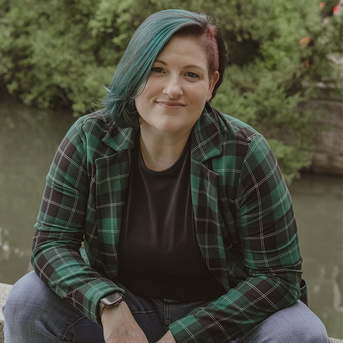 addyson tucker color portrait. addyson is genderqueer, white, small fat, and neurodivergent person who is seated on a rock wall with a stream behind them. Their elbows are resting on their bent knees as they smile gently.