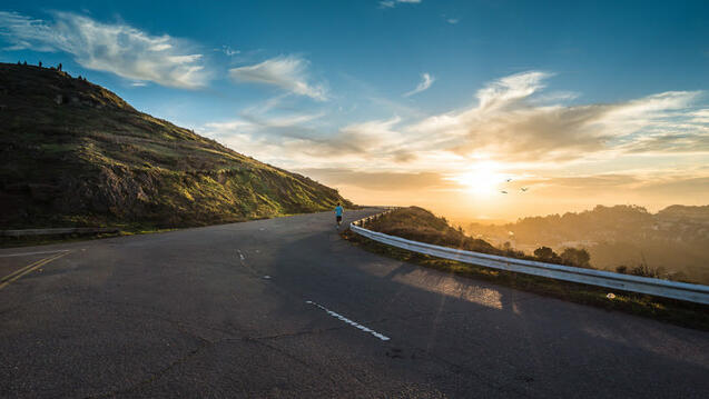 A winding mountain road with a sun setting in a blue sky.