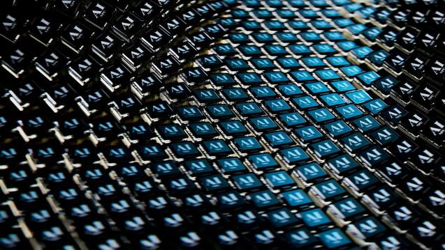 A close-up image of numerous AI chips arranged in a grid pattern, with the letters "AI" visible on each chip. The arrangement creates a dynamic, wave-like effect with alternating blue and black tones, highlighting the futuristic and high-tech nature of the components.