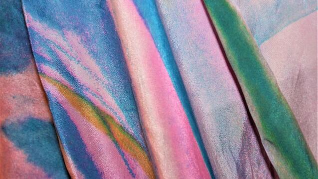 This image shows a close-up view of colorful, shimmering fabric with flowing, abstract patterns. The hues include vibrant shades of pink, blue, green, and hints of gold, creating a soft, iridescent effect. The texture suggests a smooth, silky material with subtle folds or drapes.