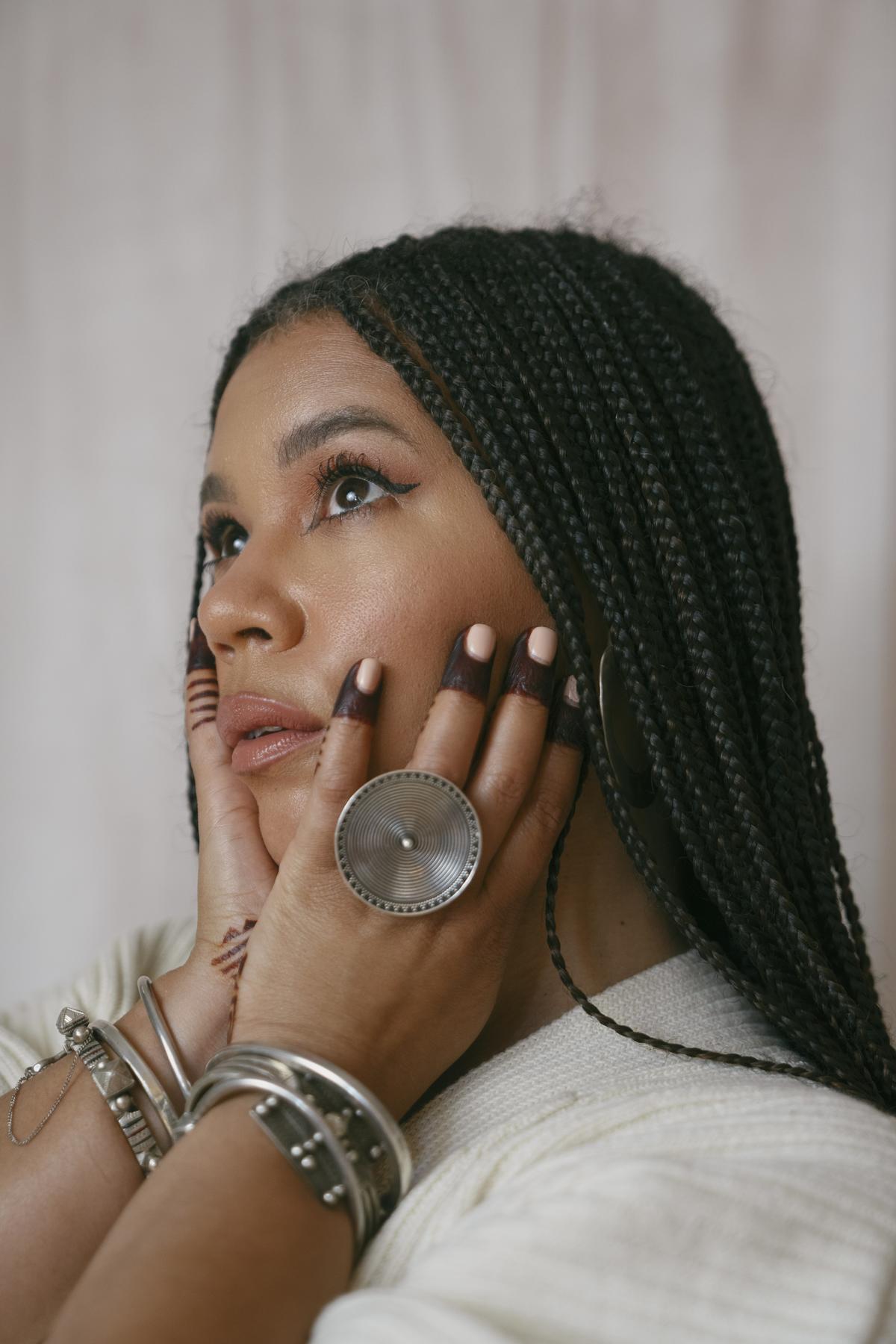 Maryam Hasnaa color portrait. Maryam is a Black woman with long, braided hair to her lower back. Her eyes are closed and her arms are raised and hands open across her chin and head.