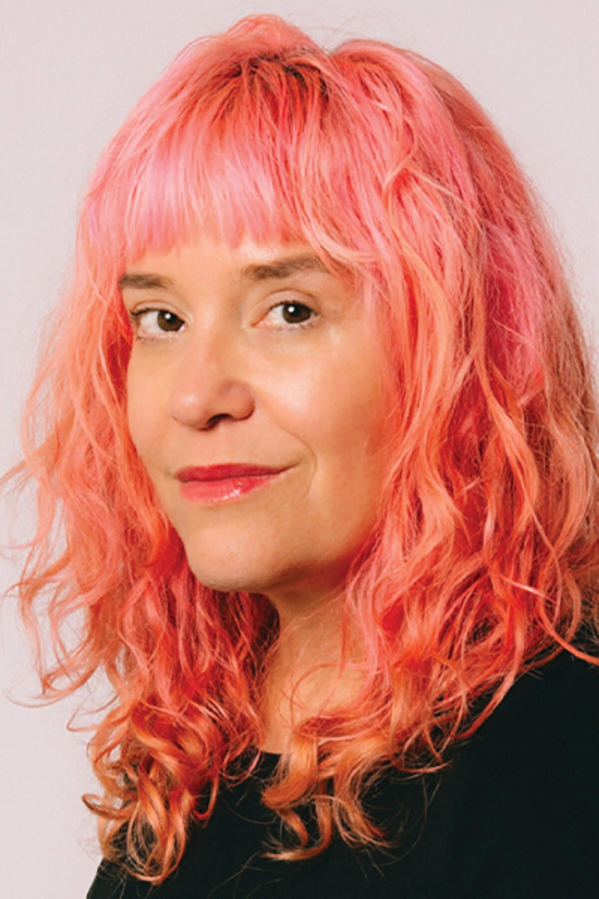 Michelle Tea color portrait. Michelle is smiling and posed against a neutral background. Michelle has soft pink wavy hair and is wearing a dark-colored top. 