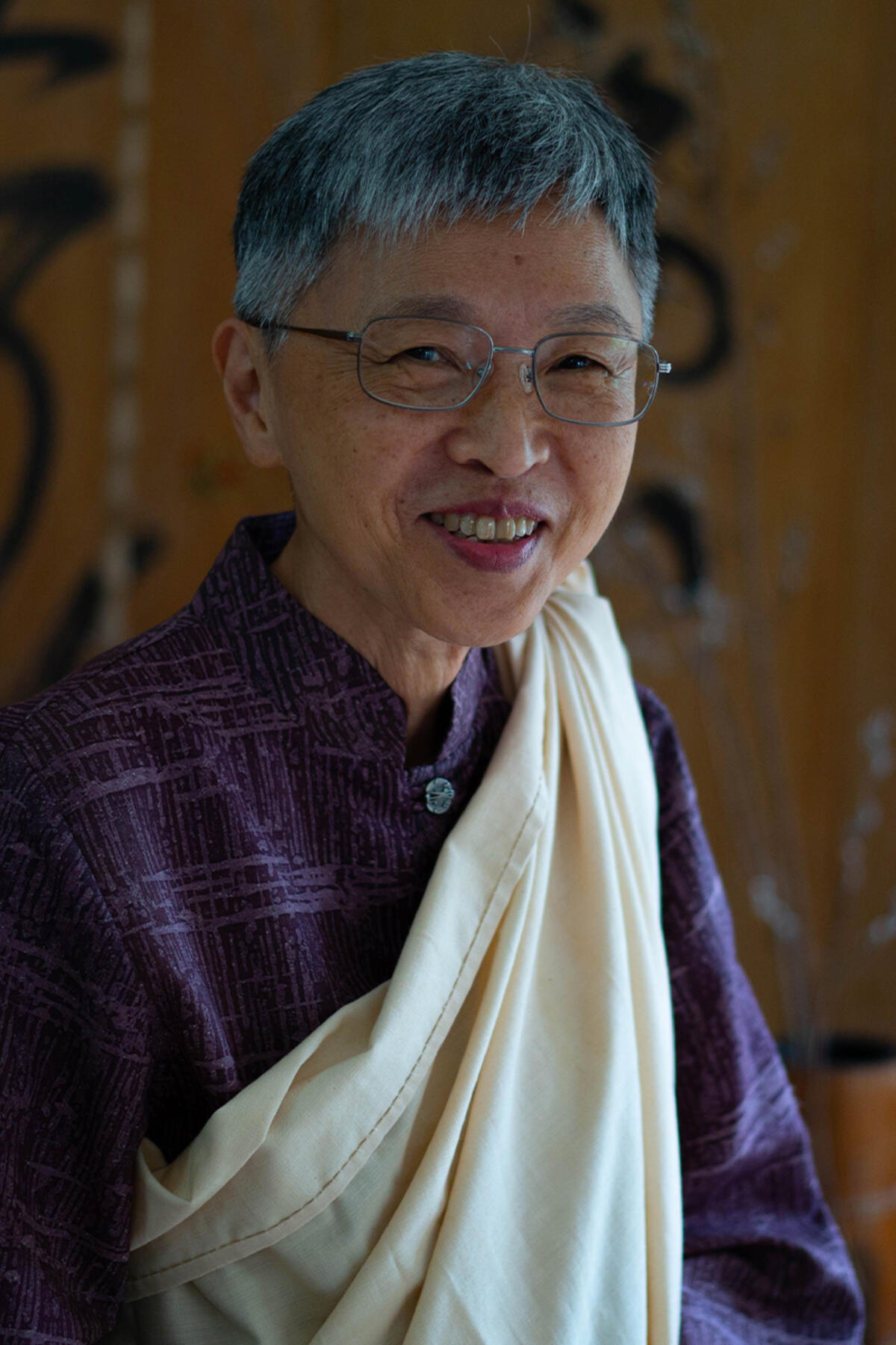 Norma Wong color portrait. Norma is Native Hawaiian and Hakka life-long resident of Hawai’i. Norma is smiling in this portrait and is posed in front of panels that have Japanese writing on them. Norma is wearing a light-colored fabric draped from one side to one shoulder and a dark colored plaid-esque printed top. 