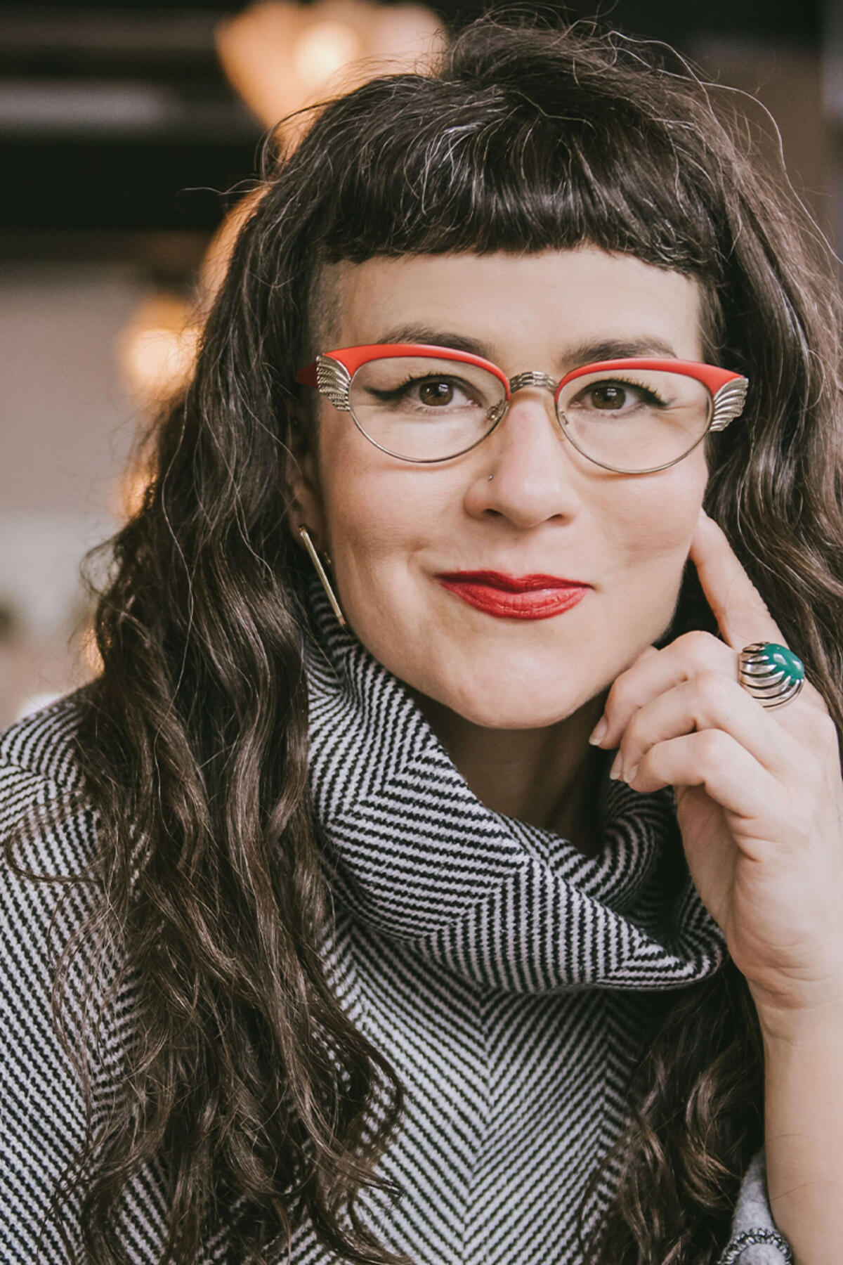 Beatriz Albina color portrait. Beatriz is a white Latina woman with long curly, brown hair that ends well past her shoulders. She is wearing red-rimmed glasses and has one hand resting on her cheek as she smiles. She is posed inside and is wearing a tweed-like top and a ring on the pointer finger that rests on her cheek.