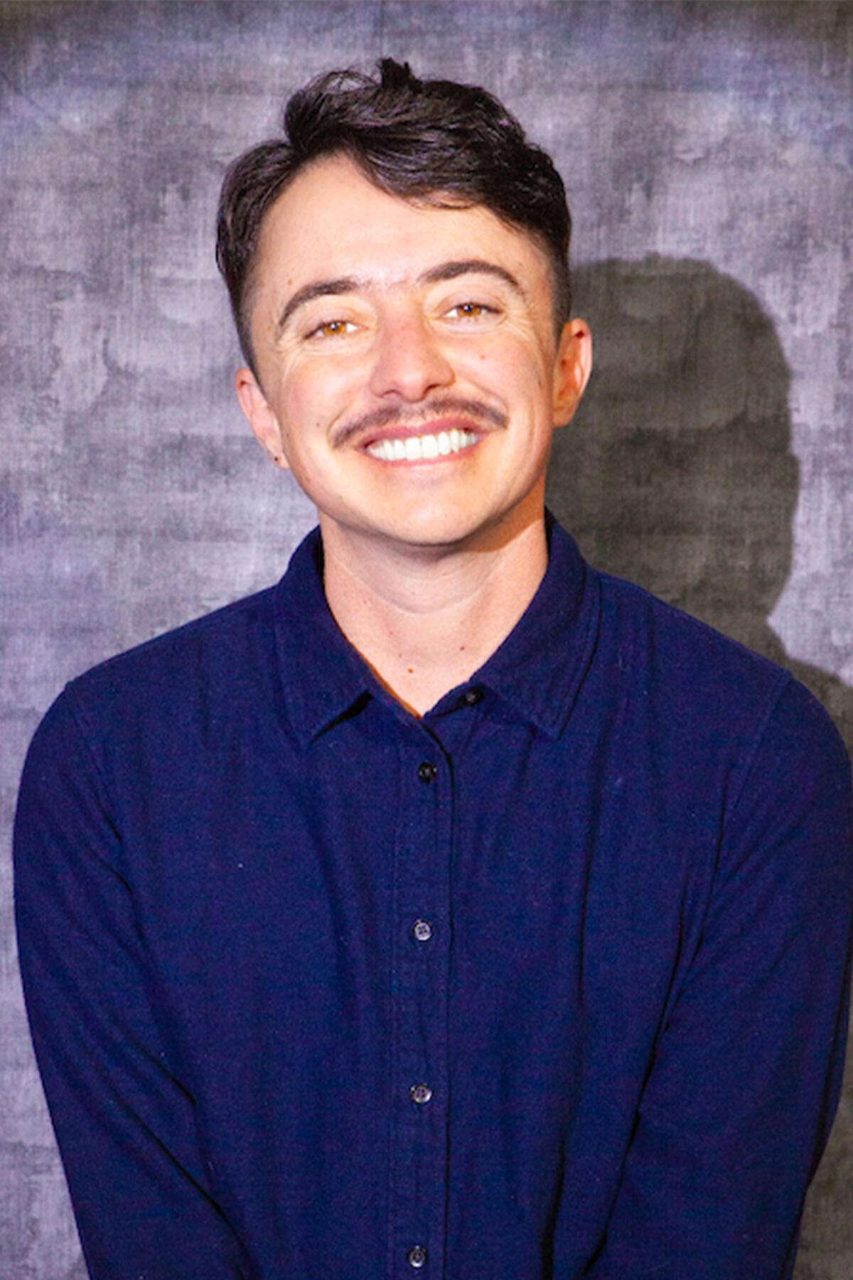 Dean Spade color portrait. Dean Spade is a white American, specifically of Jewish ethnicity; he is openly trans. He is smiling and posed in front of a light blue cloth and is seated on a stool wearing a dark-blue button-down.