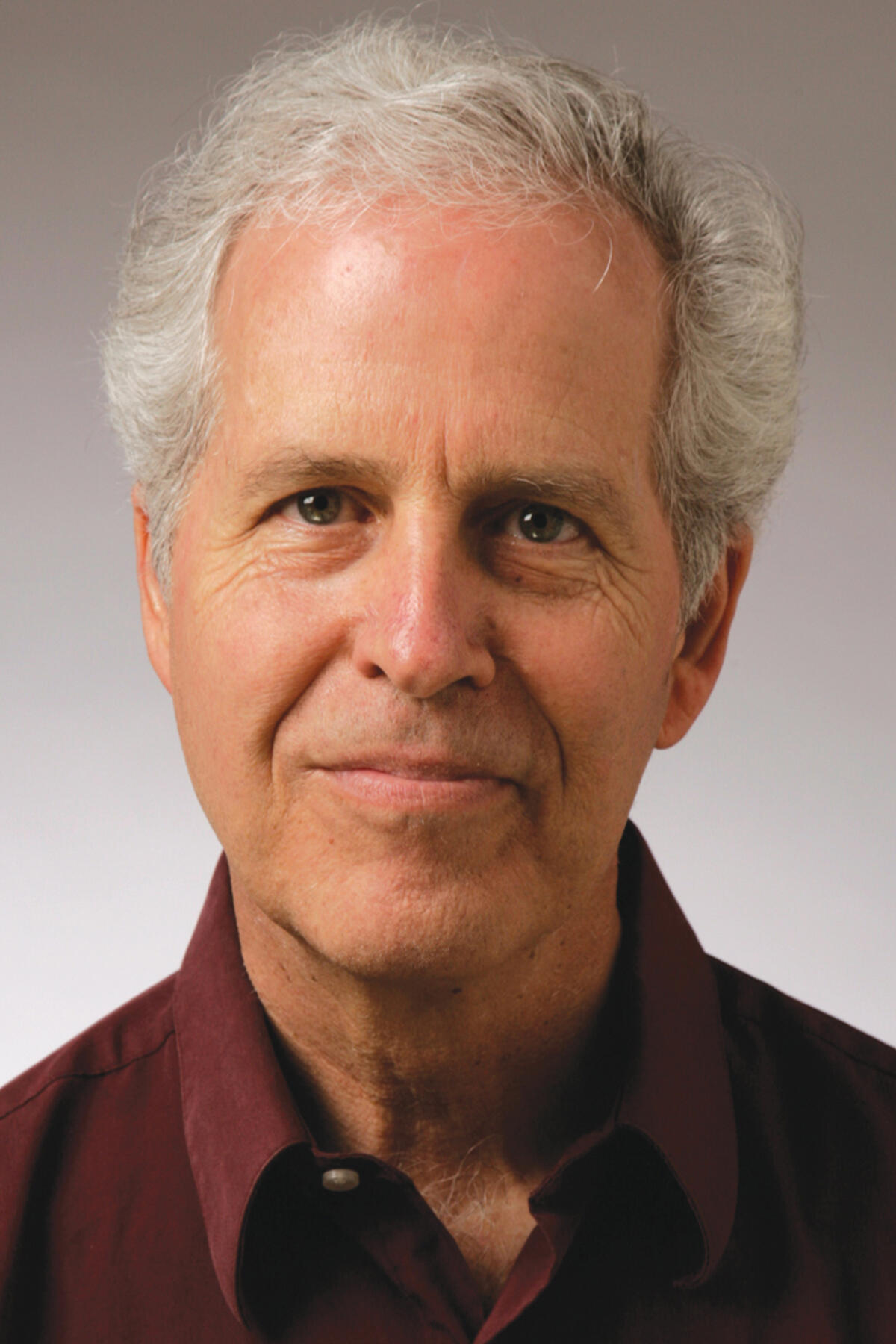 John Prendergast color portrait. John is posed in front of a neutral gradient gray background and is a white, older man with short, silver hair. He is smiling and wears a dark maroon button-down.